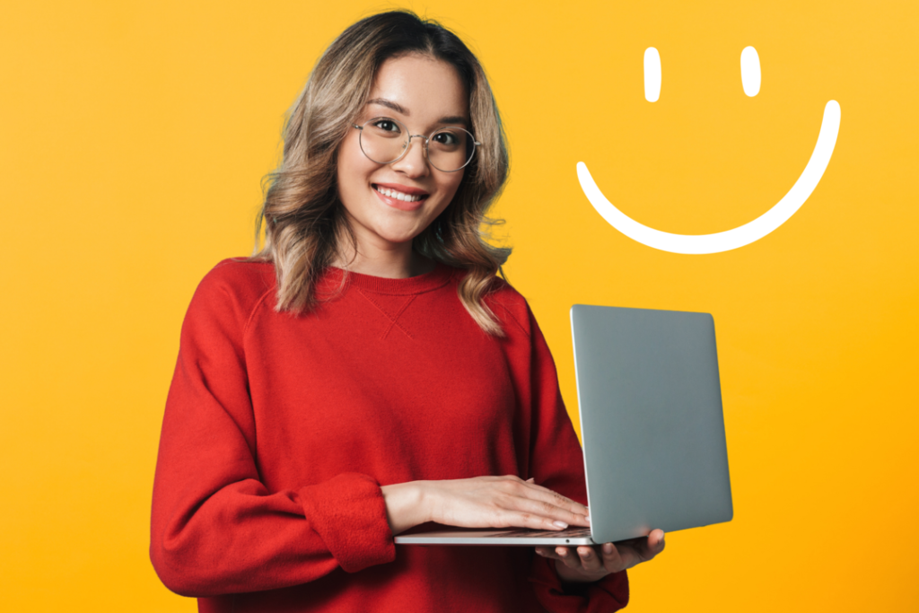Mulher jovem e sorridente, usando um óculos arredondado e uma blusa vermelha. Ela está segurando um notebook em suas mãos, em um fundo laranja.